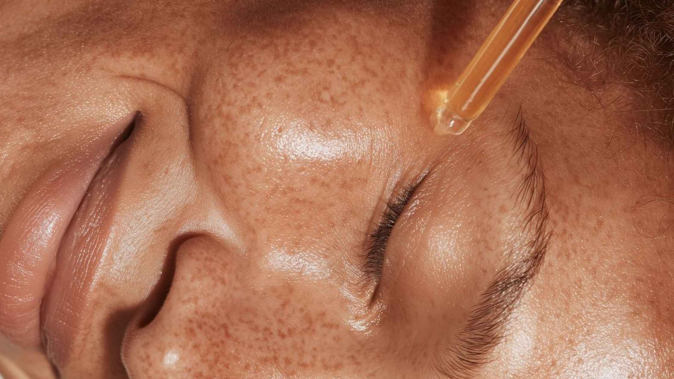 A close up of a woman smiling with a serum dropper held next to her cheek