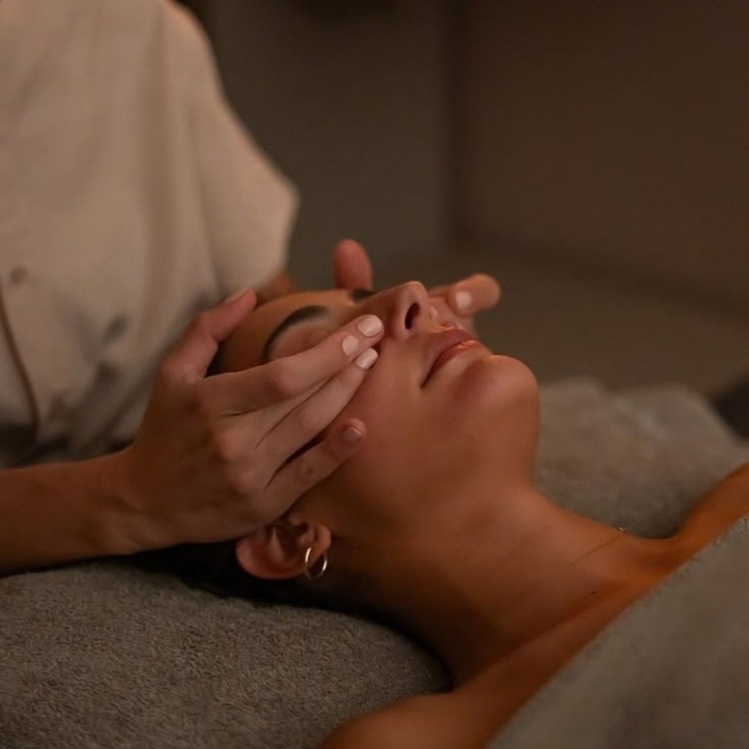A close-up lifestyle shot of a woman receiving a facial massage, lying on a gray spa towel while another person in a beige uniform gently holds her face.