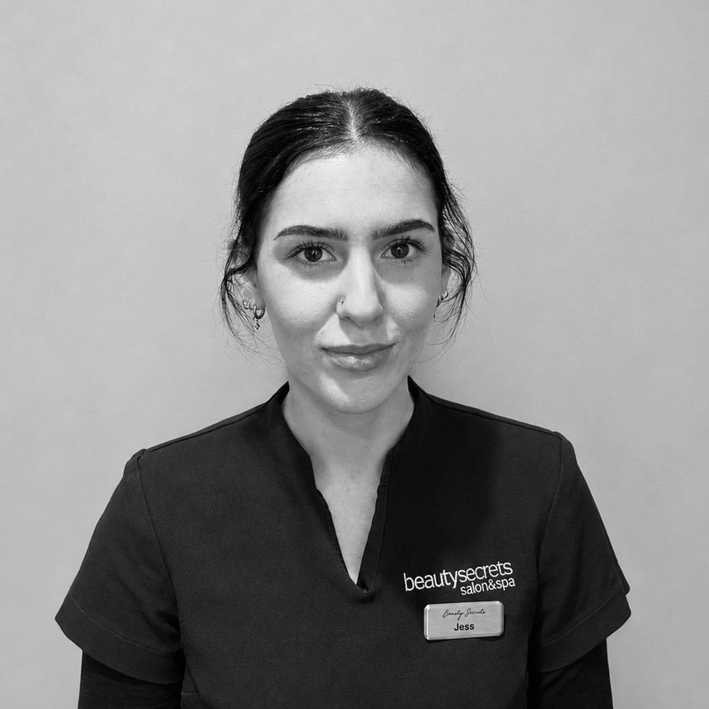 A black and white portrait of a woman wearing a dark V-neck scrub top with the logo 'beautysecrets salon&amp;spa' and a name tag reading 'Jessica'.