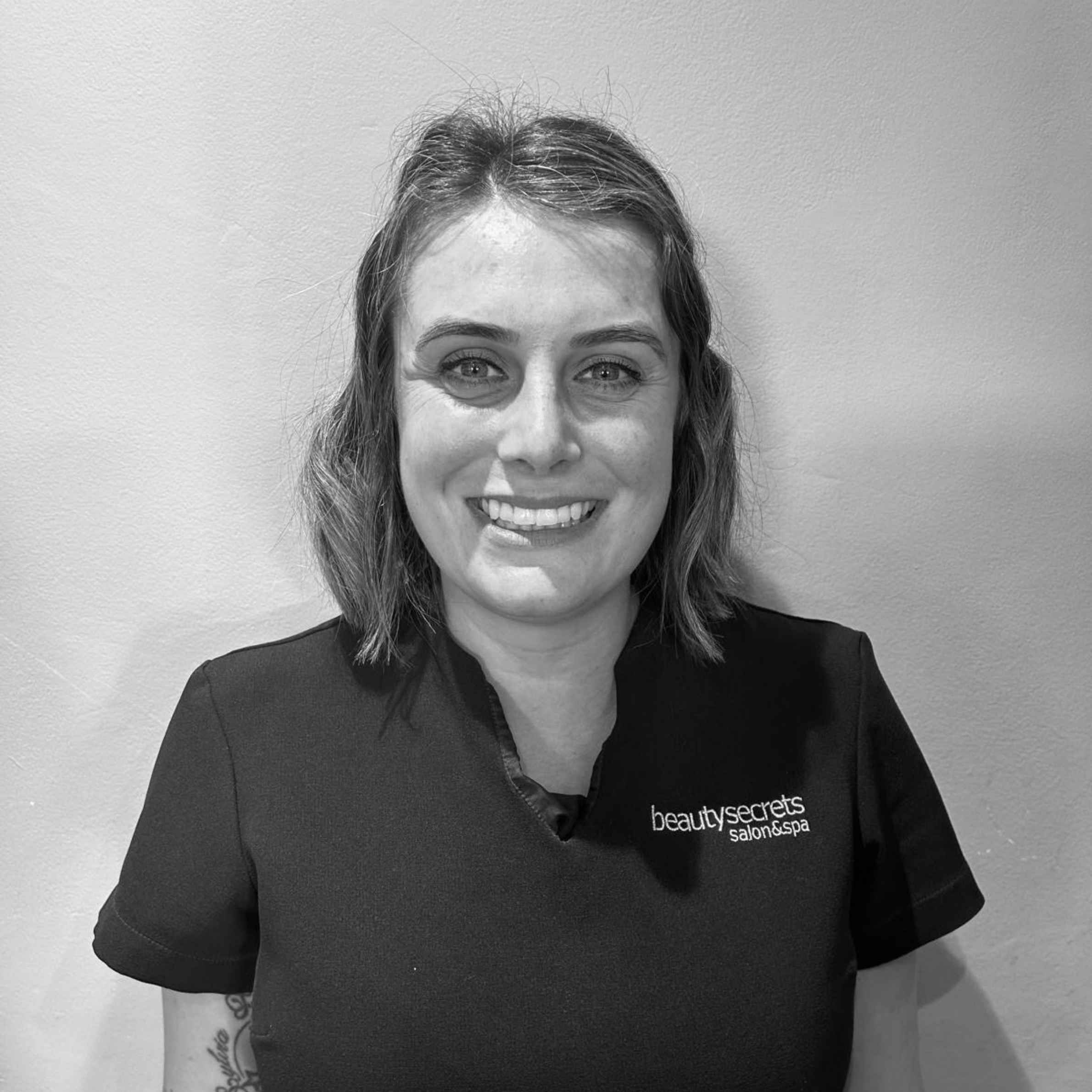 A black and white portrait of a smiling woman with shoulder-length hair, wearing a dark scrub top with the logo 'beauty secrets' on the left chest, set against a plain wall background.