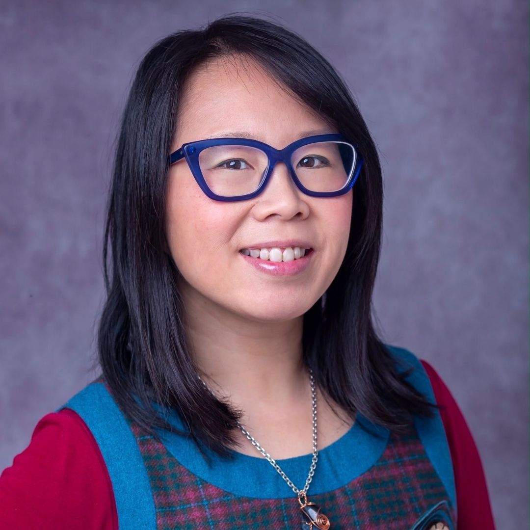 A smiling woman with shoulder-length black hair wearing blue cat-eye eyeglasses, a red long-sleeve top, and a teal-and-red plaid vest against a textured gray studio background.