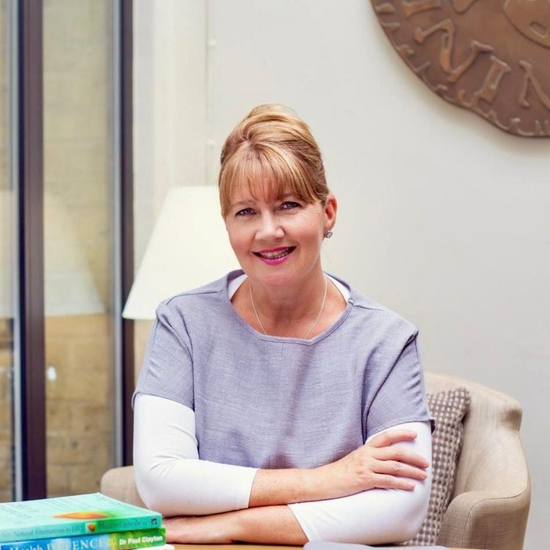 A smiling woman with blonde hair tied up sits in a beige armchair indoors, wearing a lavender-gray and white long-sleeve top. A lamp, window, and wall decor are visible in the background.