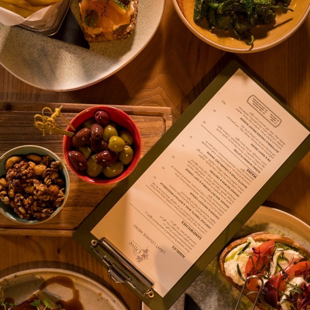 A styled food spread on a wooden board at a restaurant table, featuring sliced bread, a small bowl of olives with herbs, a gray cup, a yellow sponge, cheese wedges, and a tall black-and-orange TERRA menu or book standing behind the food. Wine glasses, a copper shaker, and a red upholstered chair are visible in the background.