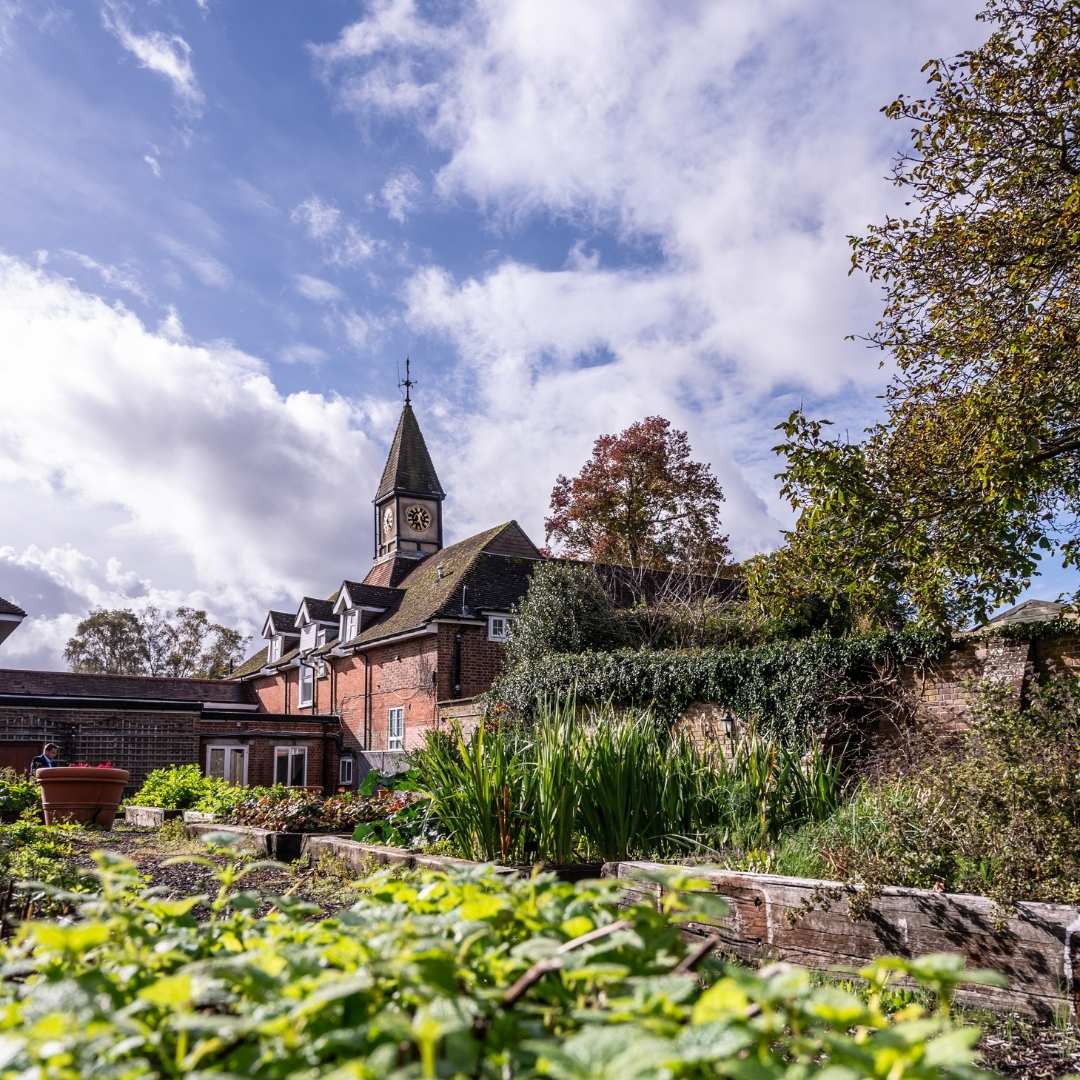 Brick building with a clock tower and garden in the foreground under a blue sky with clouds.