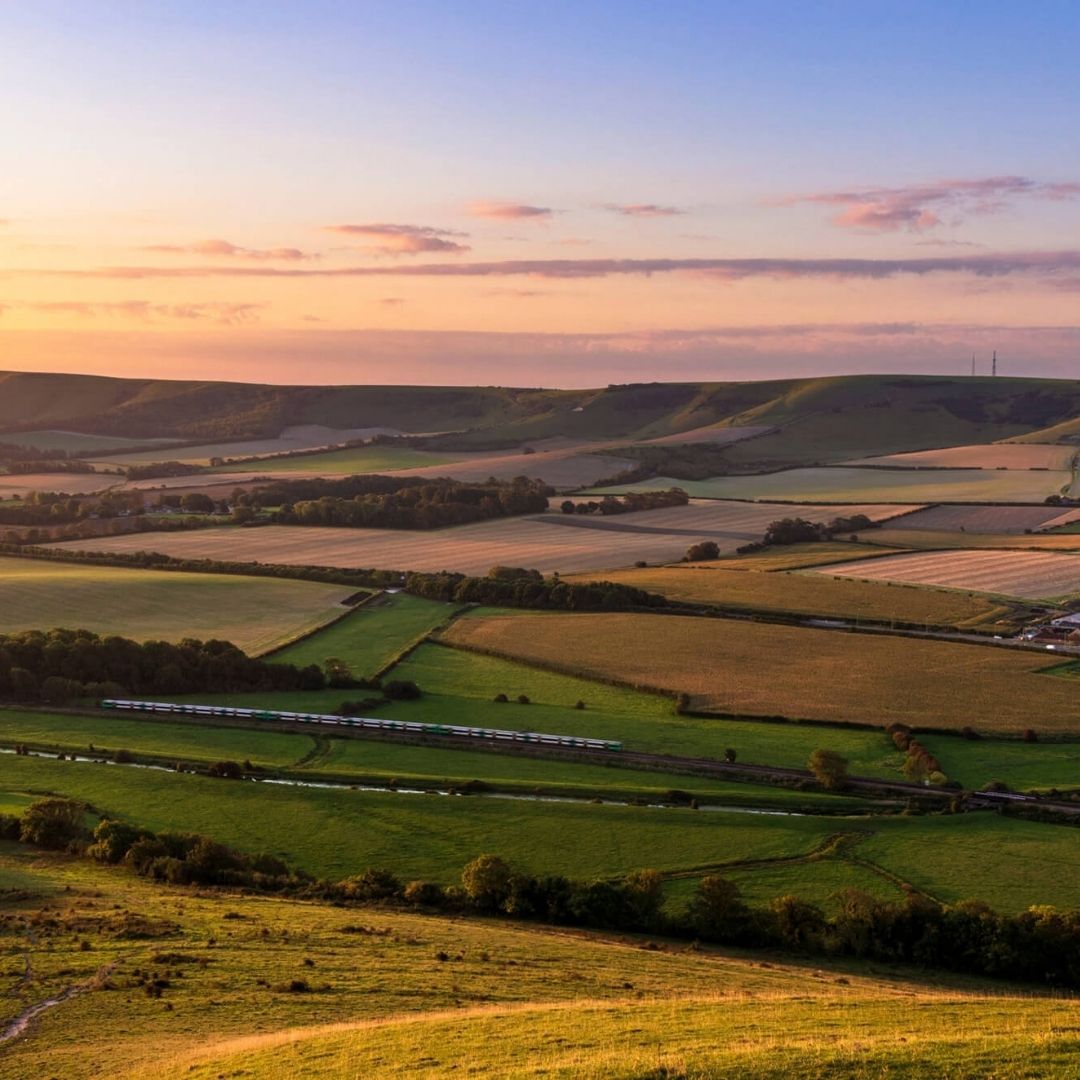 A scenic landscape photograph of a vast grassy valley with rolling hills, scattered trees, distant blue mountains, and a bright blue sky with white clouds.