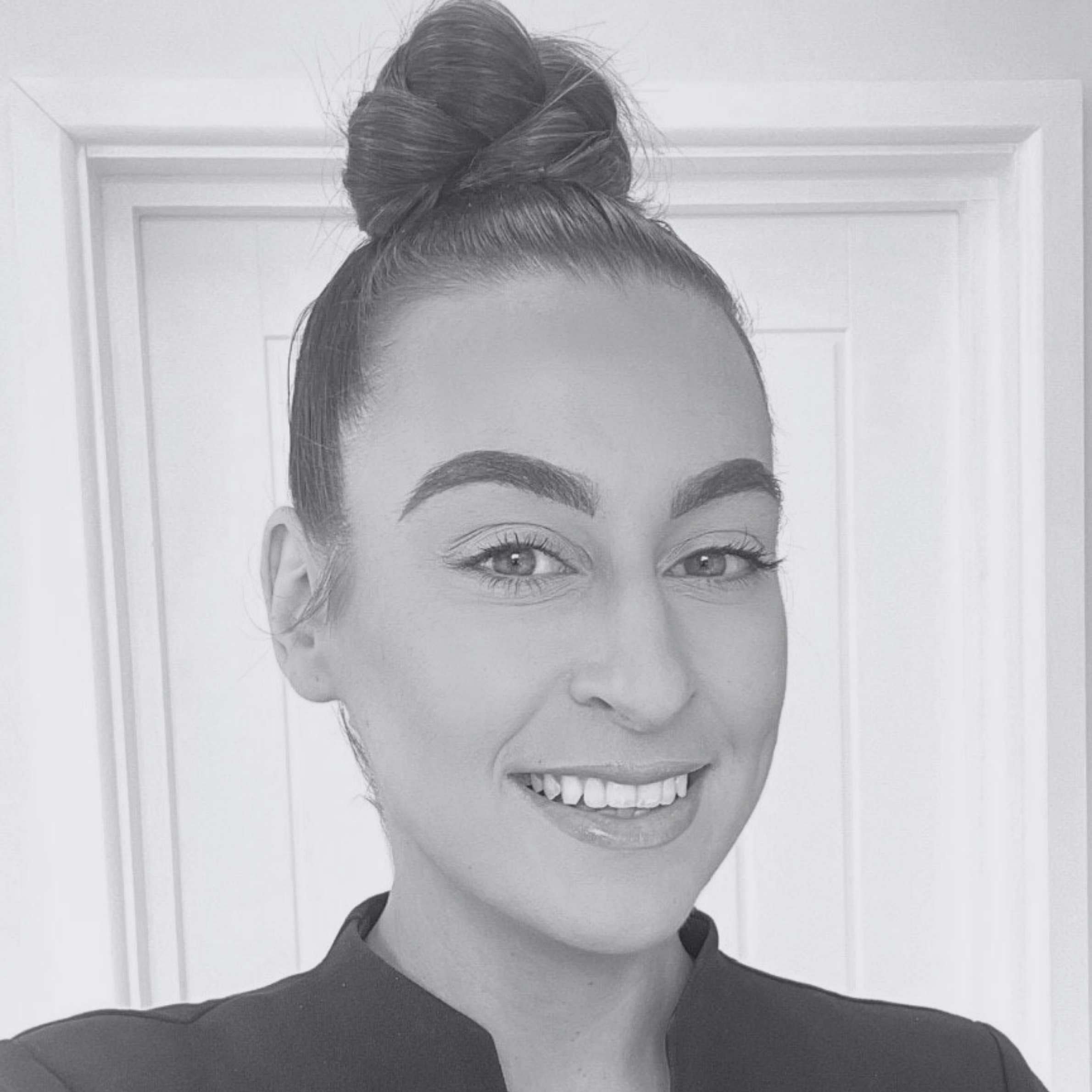 A black and white close-up portrait of a smiling woman with her hair in a high bun, wearing a dark top, set against a light-colored paneled door background.