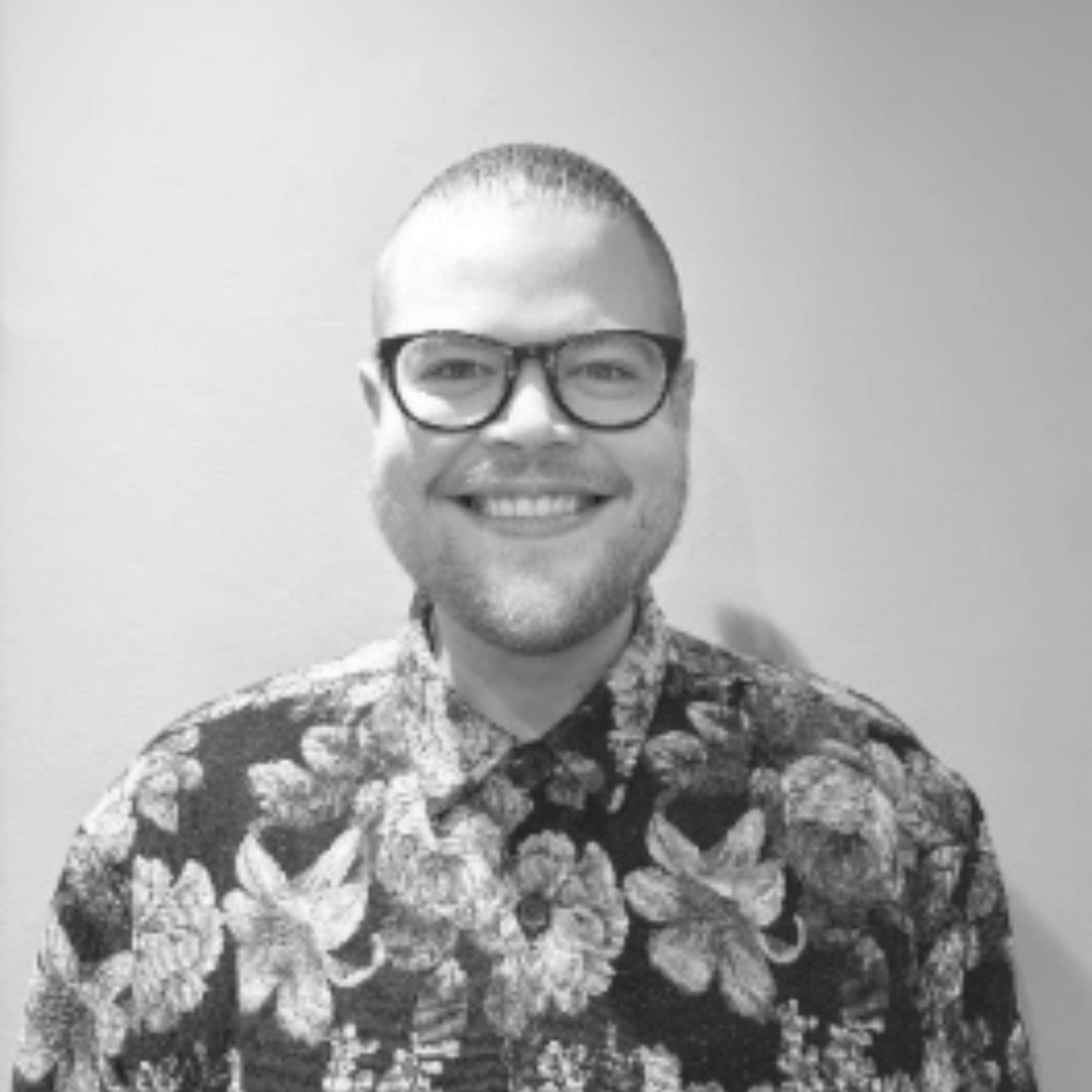 A black and white portrait of a smiling man with glasses and a patterned button-up shirt against a plain light background.