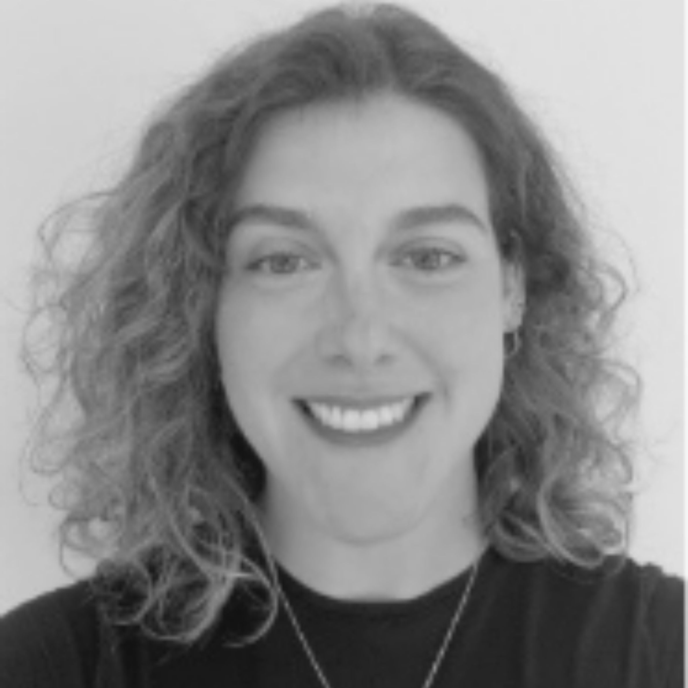 A black and white portrait of a woman with curly hair, wearing a dark satin top, set against a plain light gray wall.