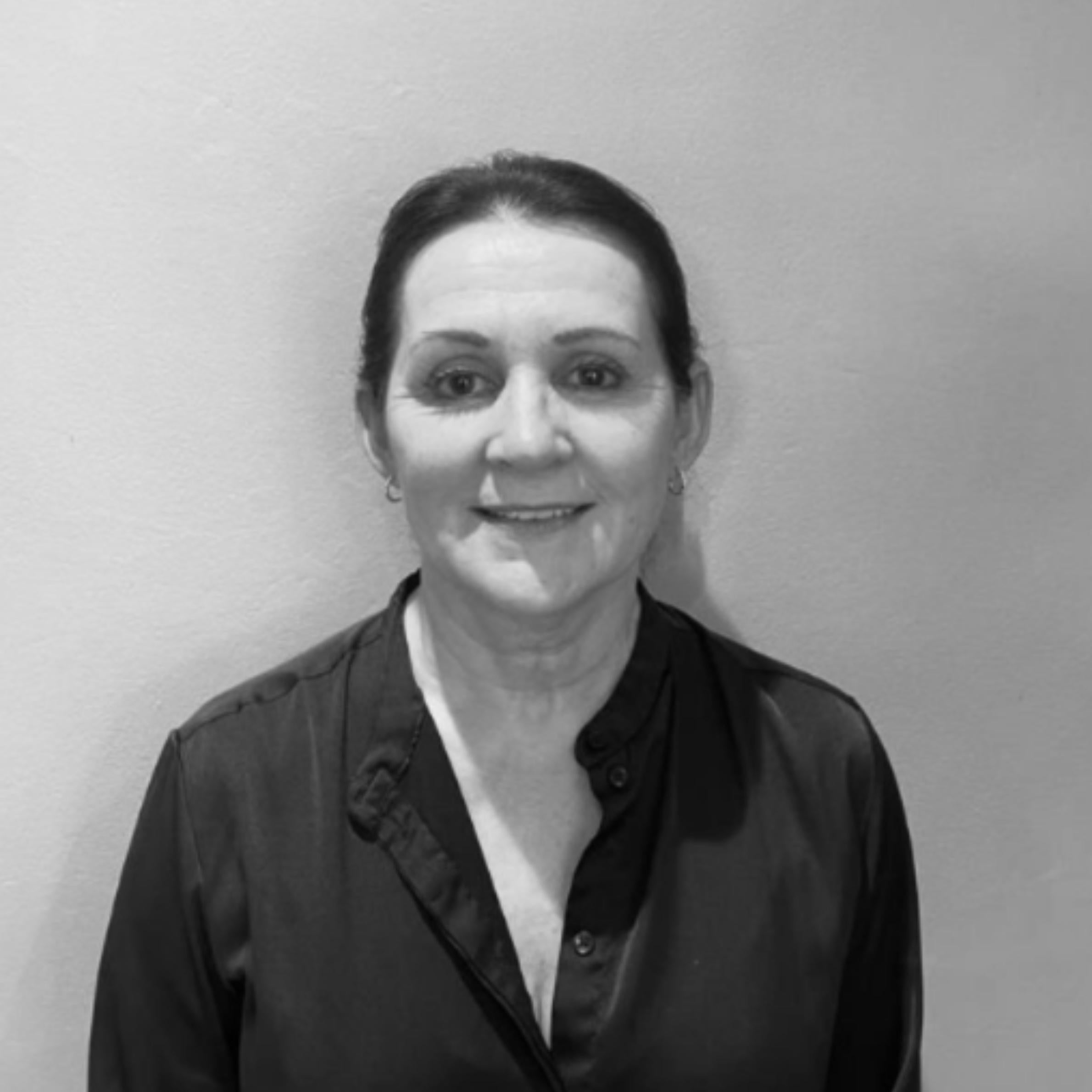 A black and white portrait of a woman with her hair pulled back, wearing a dark satin button-up blouse, set against a plain light gray wall.