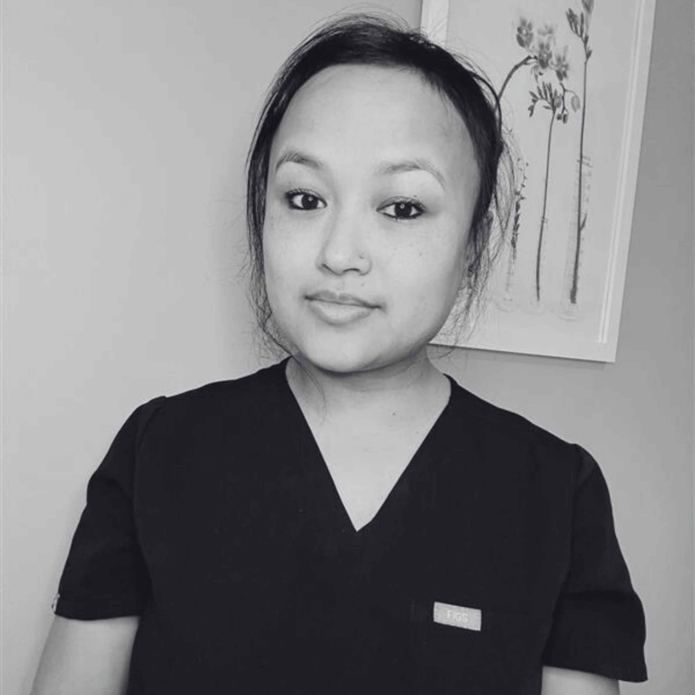 Person wearing a black scrub top with a small logo, standing against a light-colored wall with a framed picture.