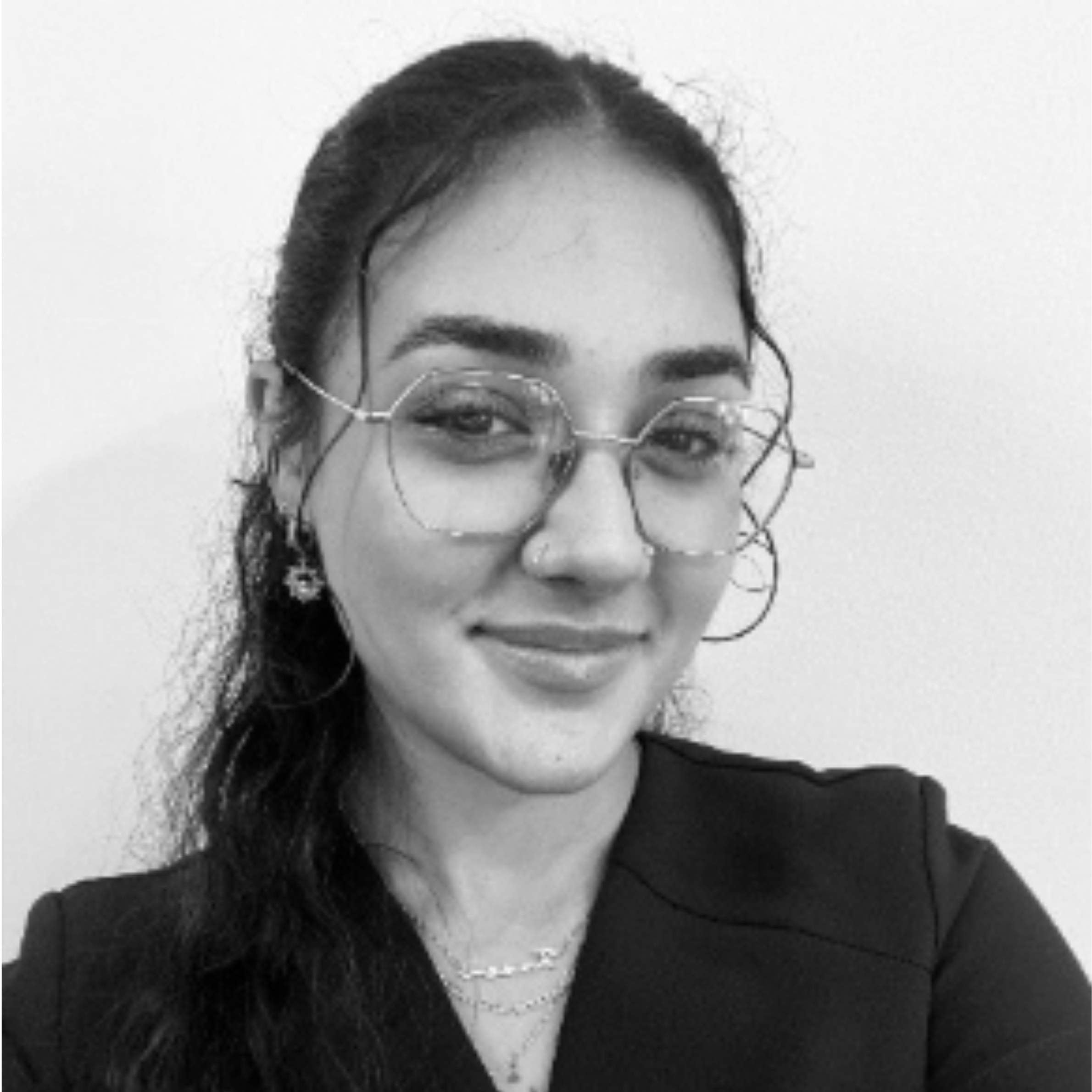 A black and white portrait of a woman with long dark hair, wearing round wire-frame eyeglasses, a nose ring, layered necklaces, and a dark blazer.